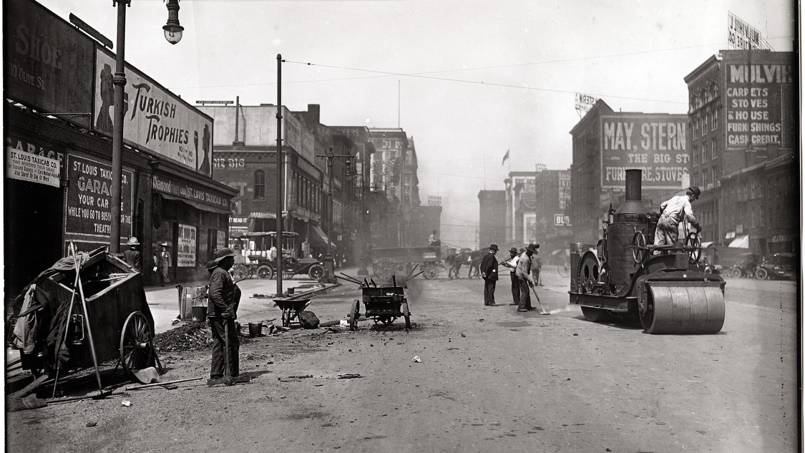 Horizontal, black and white photograph showing workers repaving 12th Street, now Tucker Boulevard, just south of Chestnut. Workers are using a large roller to smooth the asphalt, as well as a variety of smaller tools and