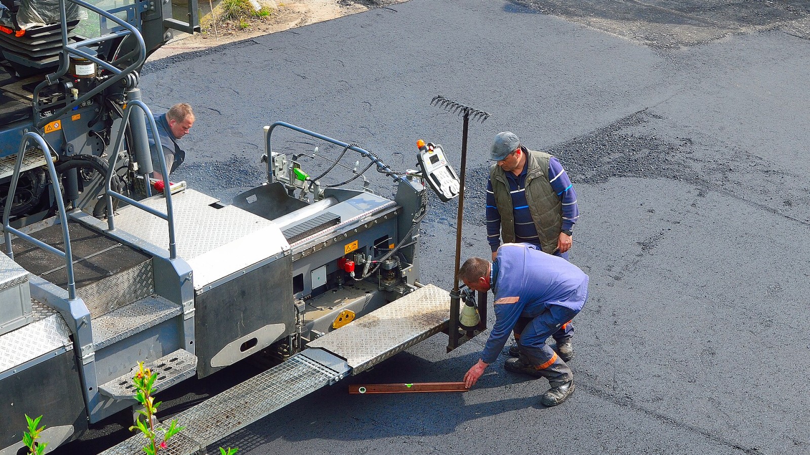 Construction workers examine new road surface with a spirit level. On the left side: An asphalt paver, Vögele Super 1800-3i.