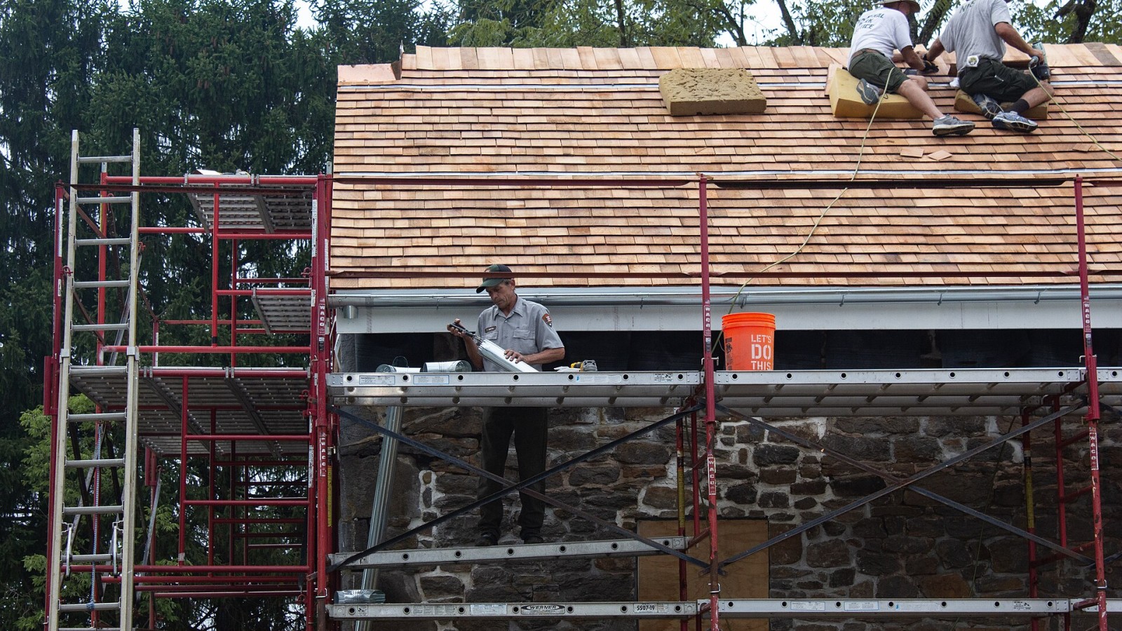 Close up image of the rear roof of the Warfield house. There is scaffolding on the left and and in the foreground. Two workers kneel on the wooden roof installing shingles at the top right corner of the image. Another st