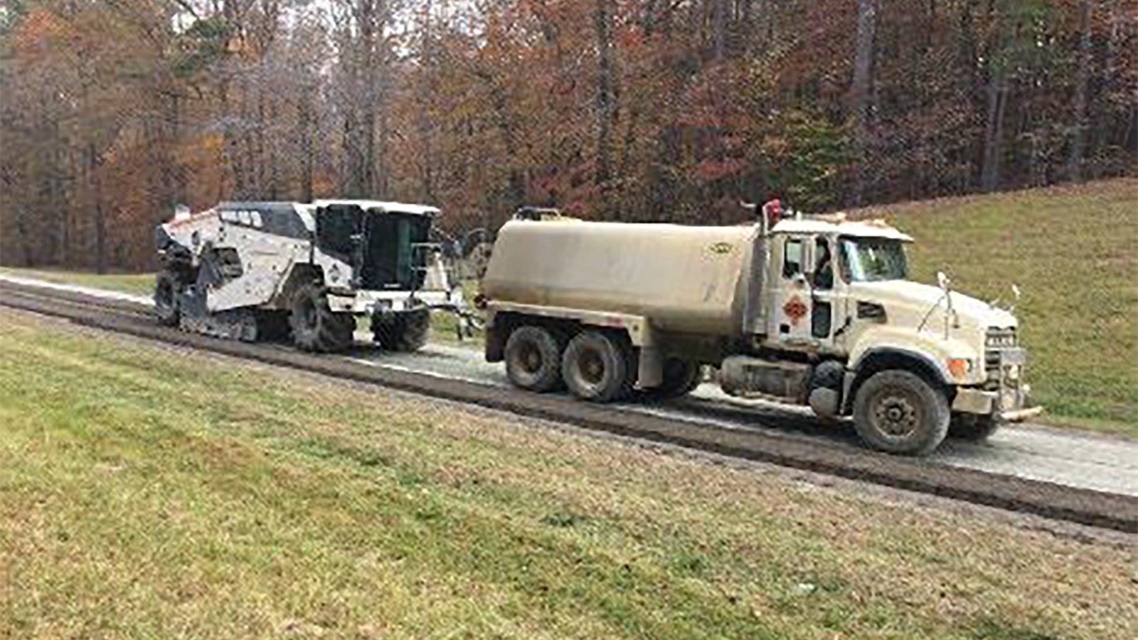 On a scraped road a reclaimer vehicle with a low, but large cab, large wheels and metal sides sits behind a water tanker truck. On a scraped Parkway, a reclaimer vehicle sits behind a water truck for mixing concrete. Key