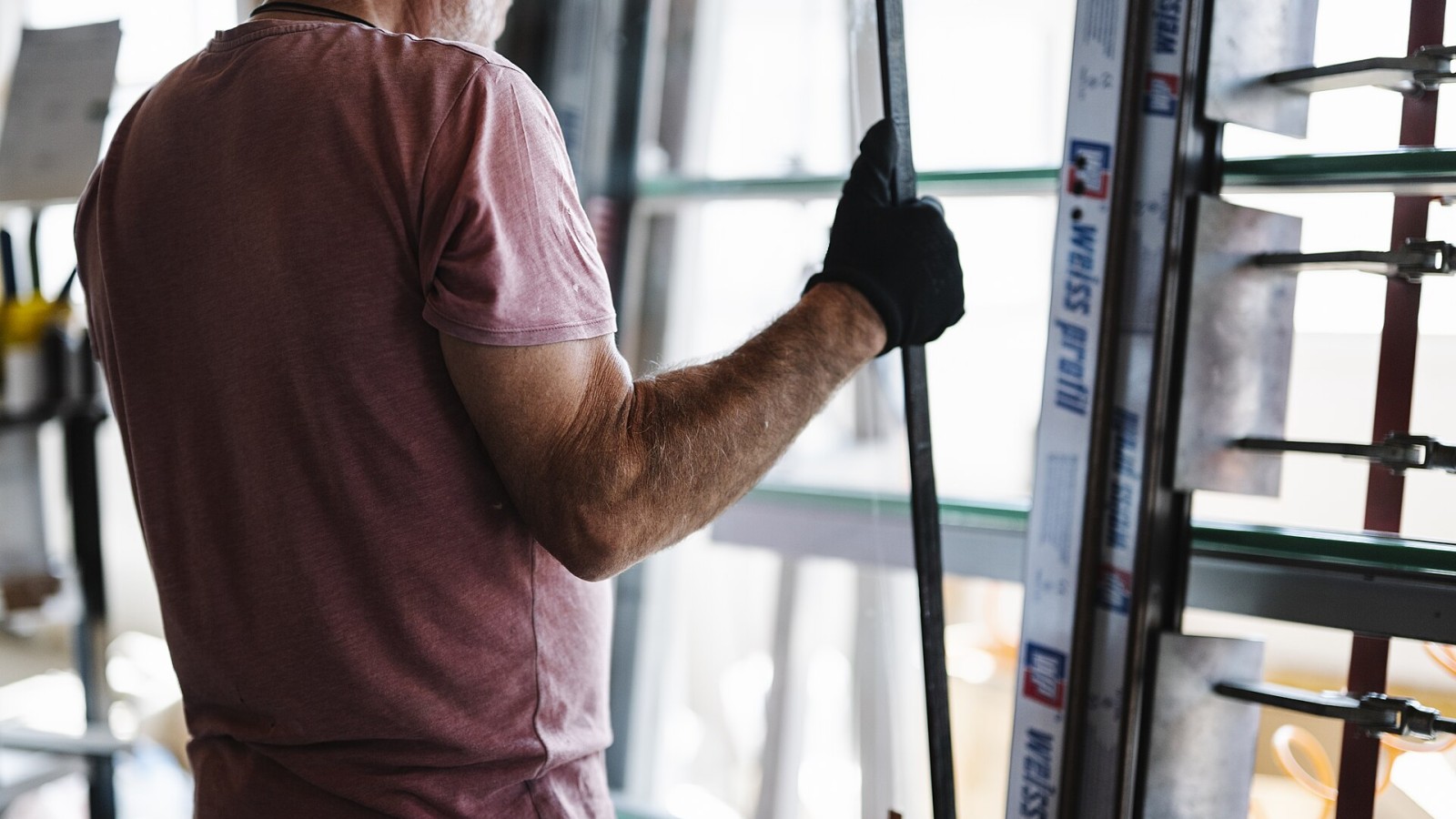 A person stands inside a building under construction, holding a glass panel with one hand. Tools and equipment are nearby. The scene shows part of the glass installation process.