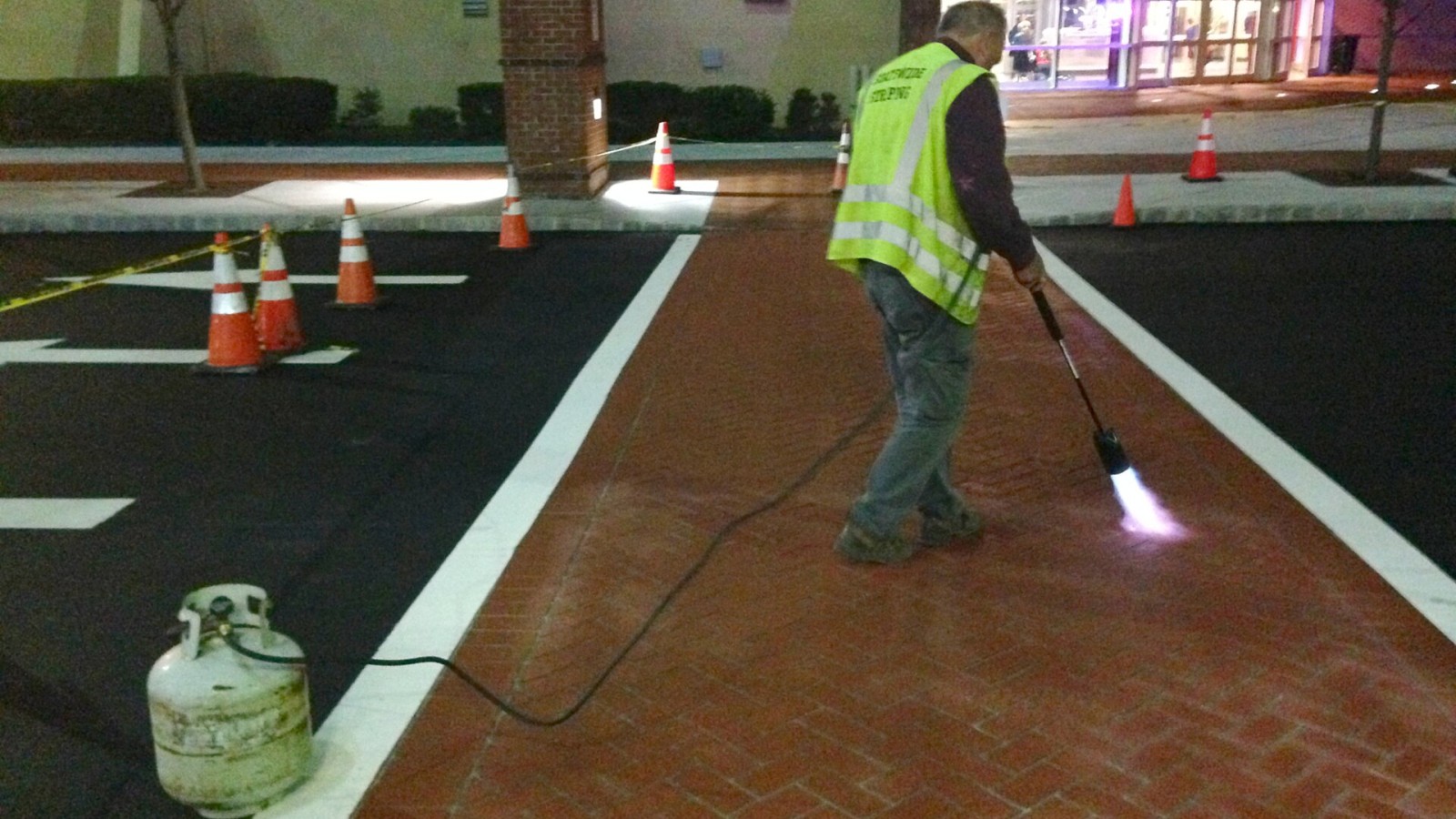 A worker uses propane torch to speed cure the colored polymer cement slurry that has been applied over a brick stencil to create a decorative crosswalk. The steps of crosswalk construction include spraying a layer of gra