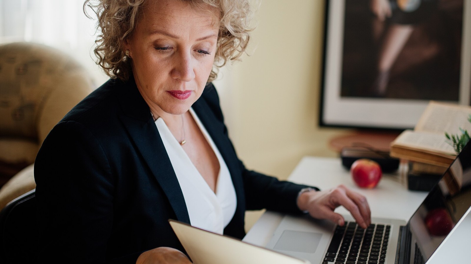 A focused professional woman sits at her desk, engaging with a laptop and reviewing documents. Natural light filters through the window, enhancing the serene workspace atmosphere.