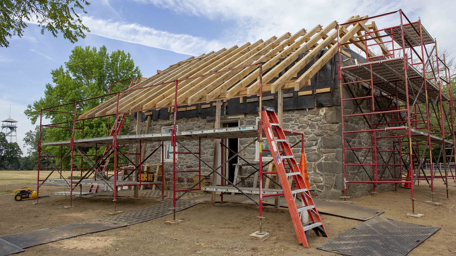 Angled photo of the front and right side of the Warfield house. The bare wood rafters are exposed on the top of the stone walls. There is a large set of red metal scaffolding on the right side of the building and a short