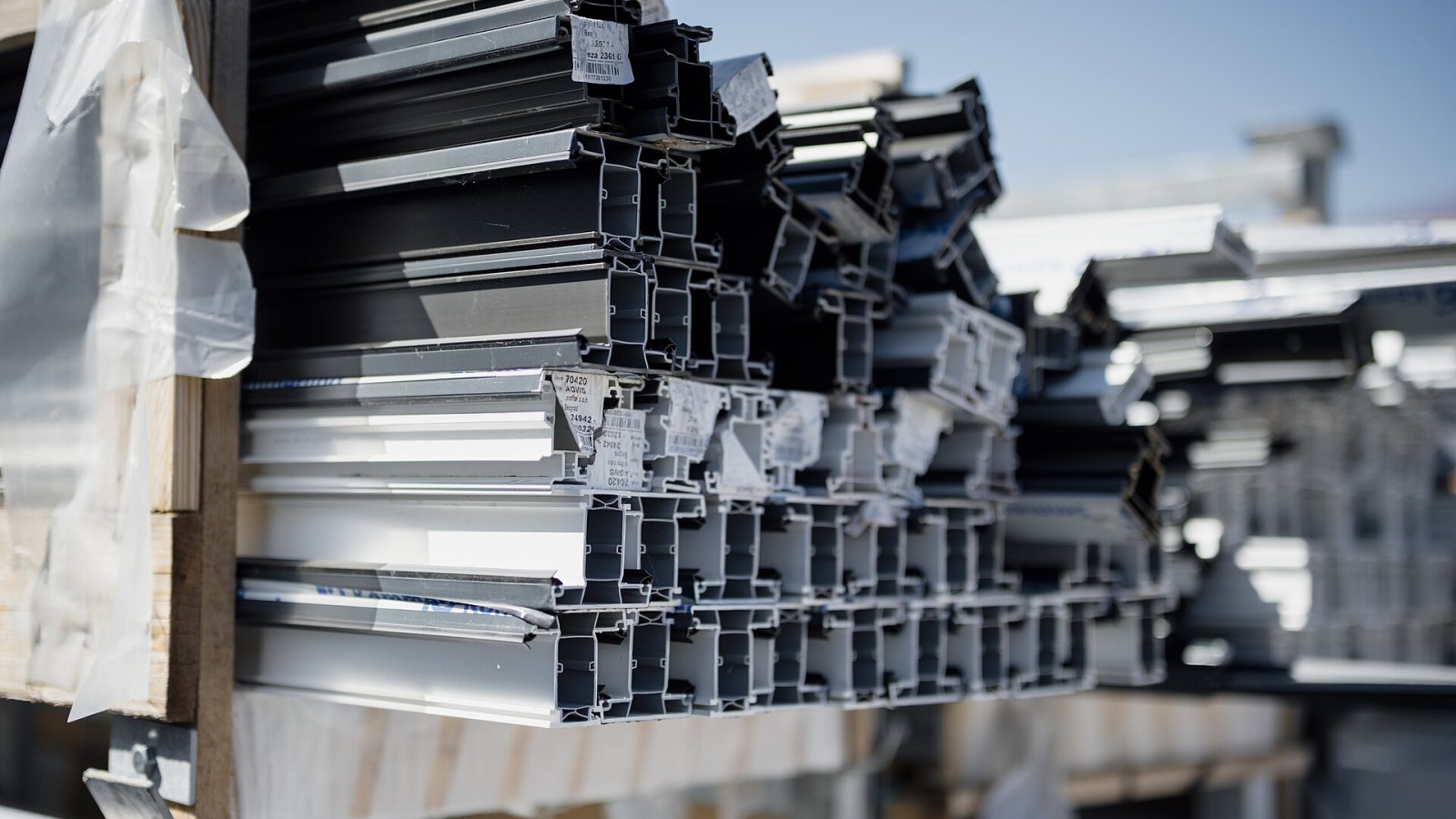Workers prepare for a building project by organizing metal frames at a construction site. The frames rest on wooden pallets in the sun.