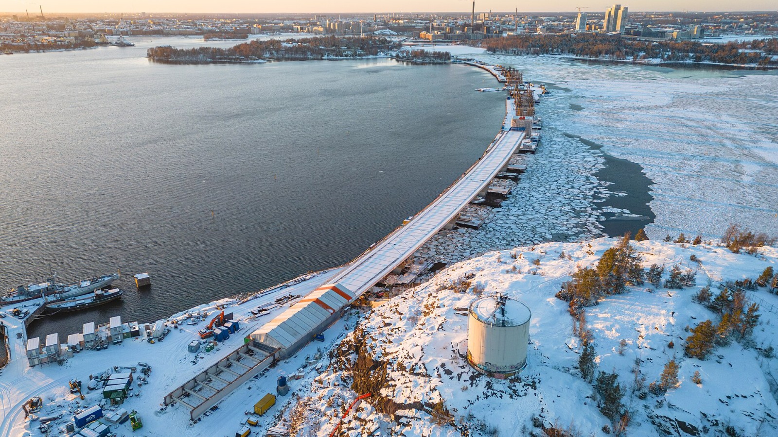 Aerial view of the construction site of Kruunuvuori Bridge connecting Kruunuvuorenranta and Korkeasaari. Old Toluene tank on top of Kruunuvuori cliffs. Buildings of Helsinki City Centre and Hanasaari Power Plant in the b