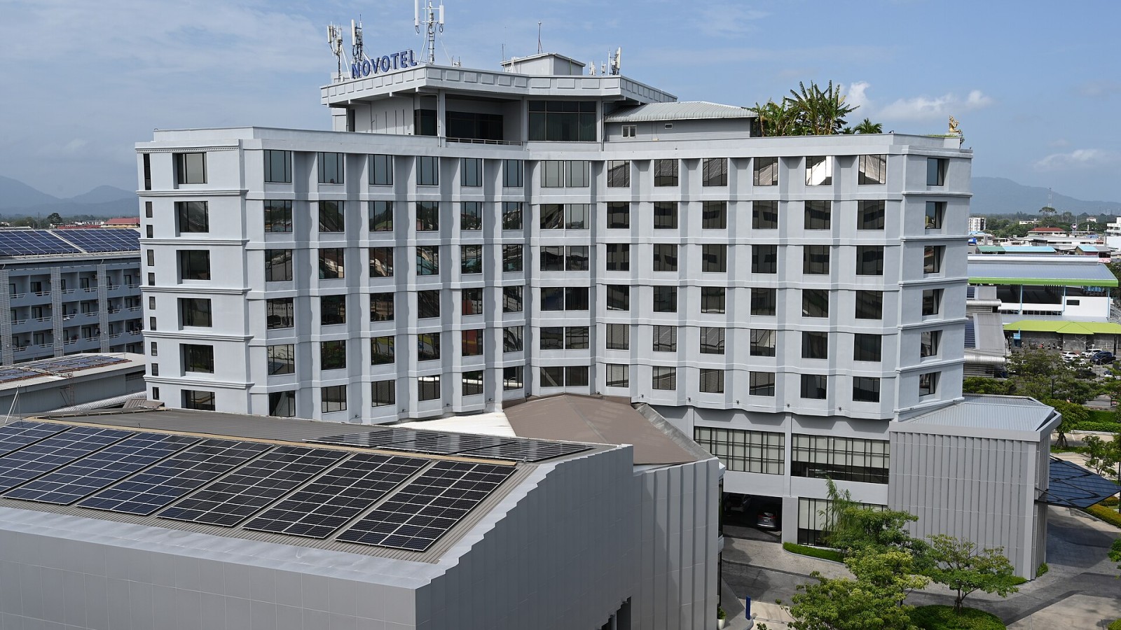 This photo shows a modern hotel or resort building, which appears to be a NOVOTEL property based on the signage visible on the roof. The structure features a multi-story white building with numerous windows and balconies